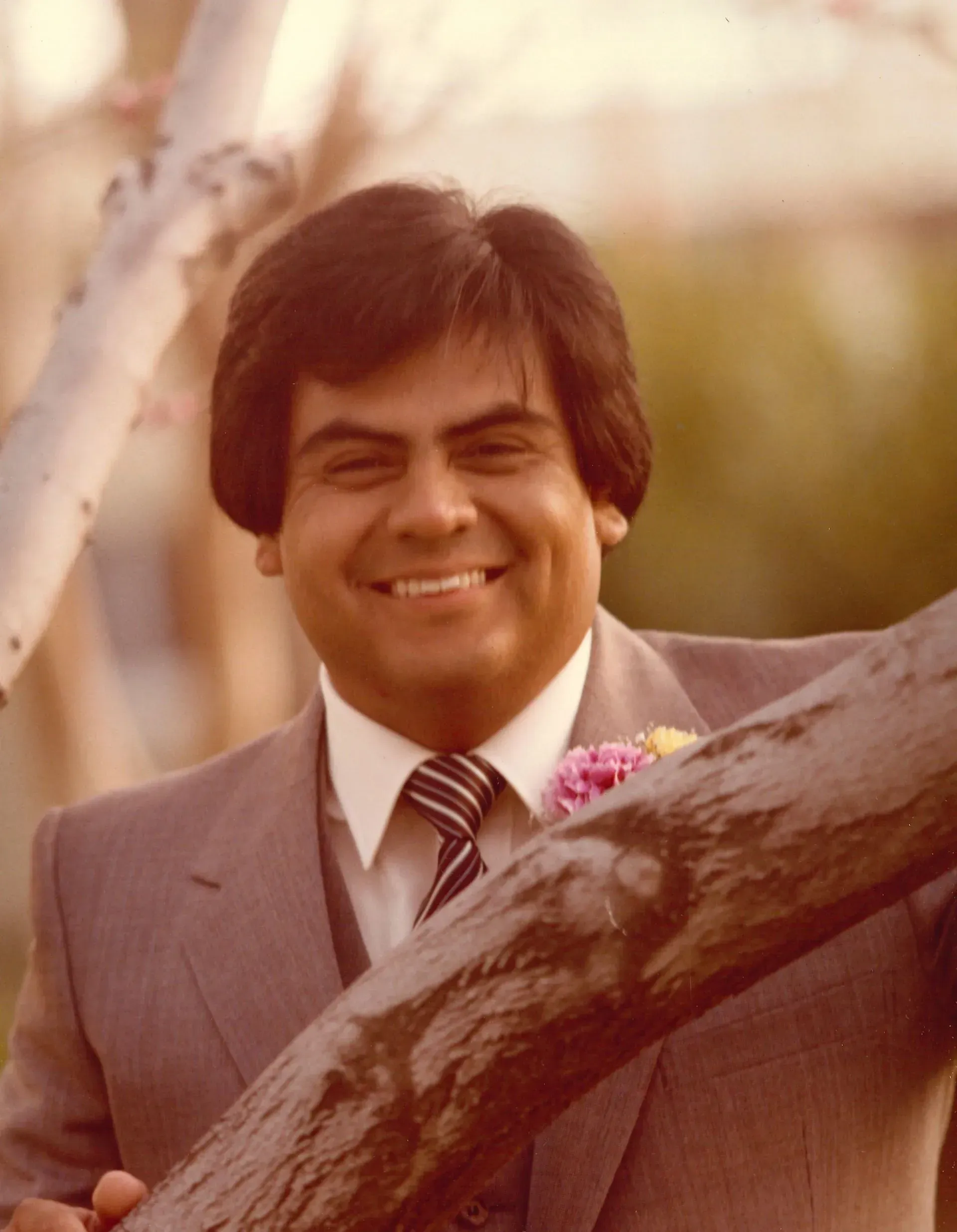 Smiling man in a gray suit stands outdoors beside a tree branch