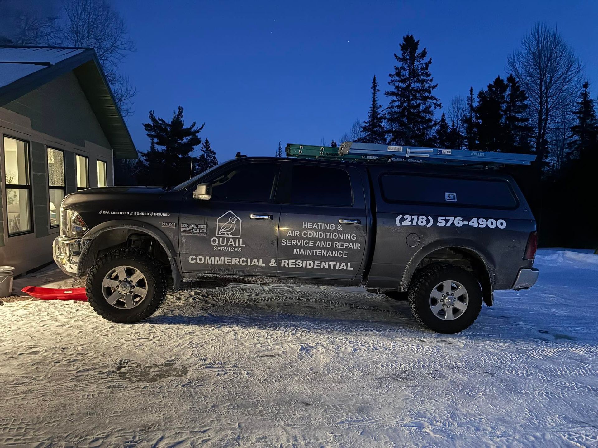 Black service SUV parked in snowy driveway at dusk beside a lit house, with company lettering and phone number on the side