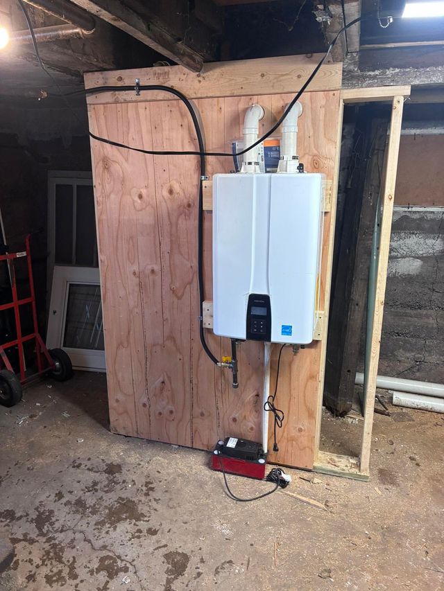 White boiler mounted on plywood wall in unfinished basement, with exposed pipes and wires.
