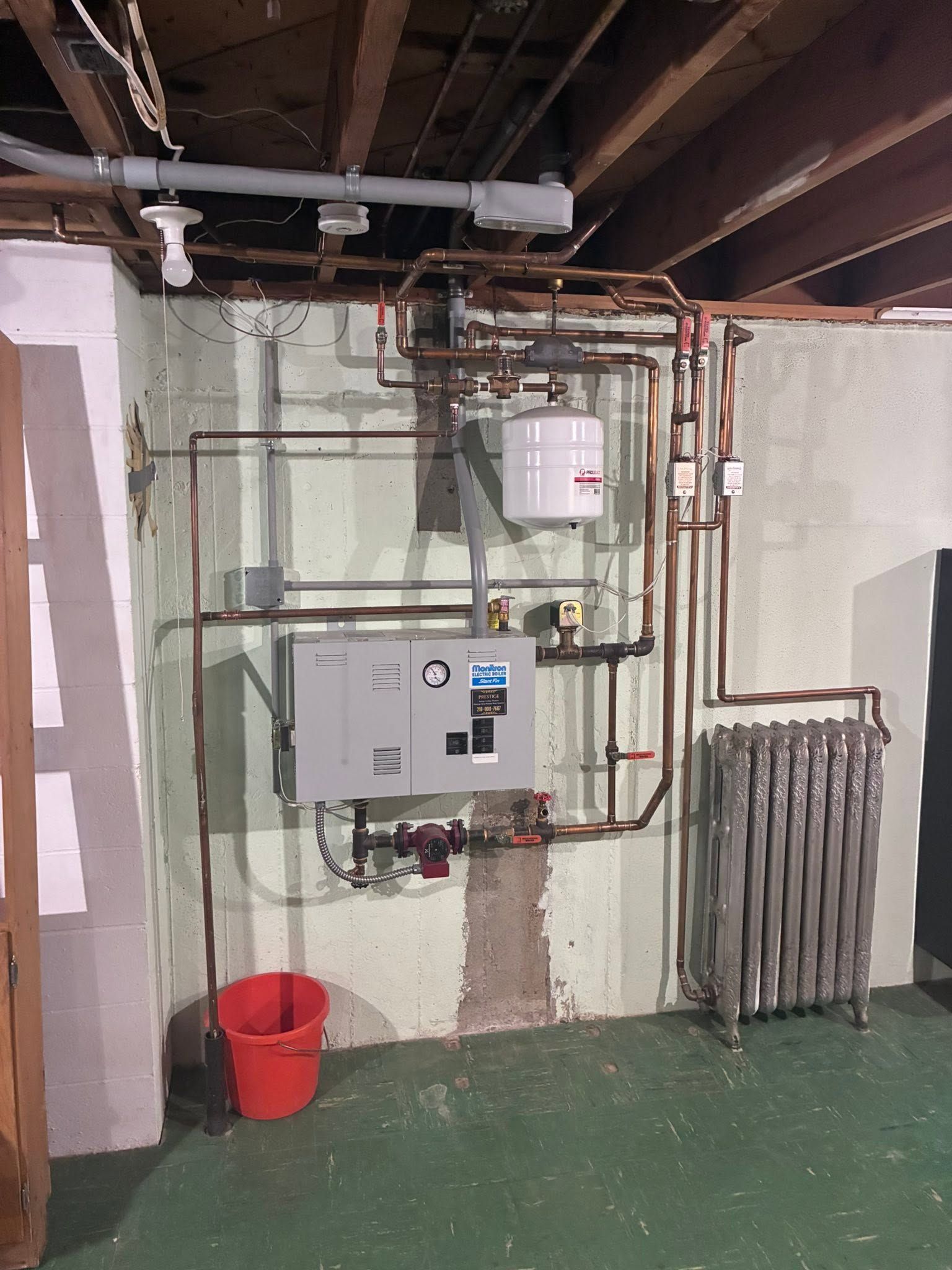 Basement utility room with electrical panels, exposed pipes, a red bucket, and a radiator on the right.