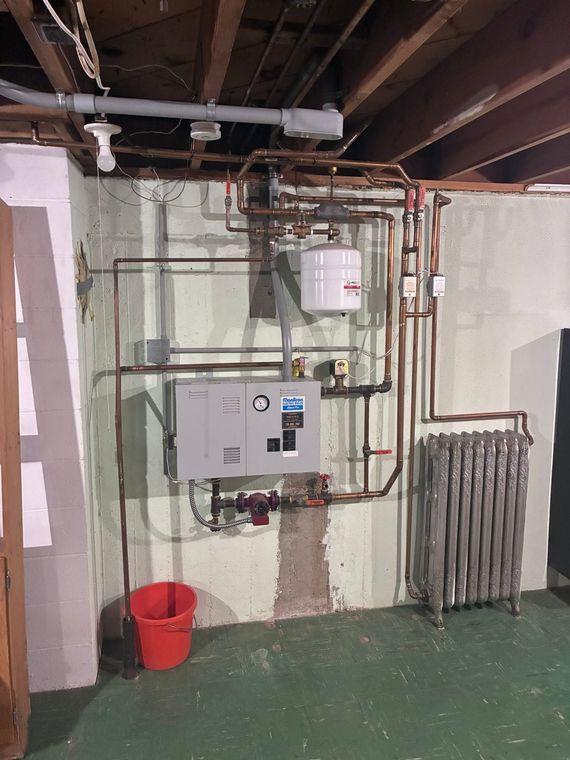 Basement utility room with electrical panel, water heater, pipes, radiator, and red bucket on green floor
