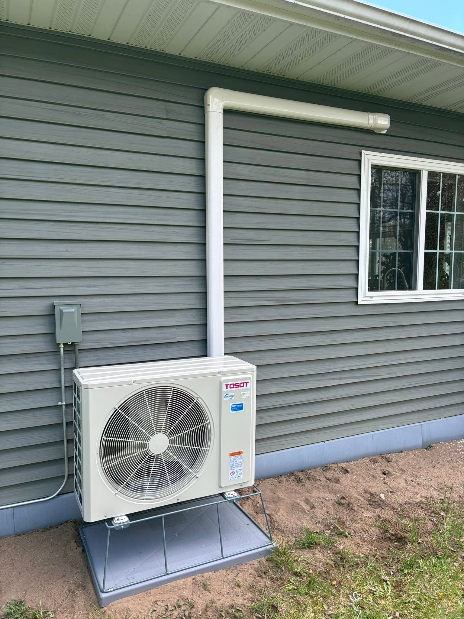 Outdoor heat pump unit mounted beside a gray house with white piping and a window