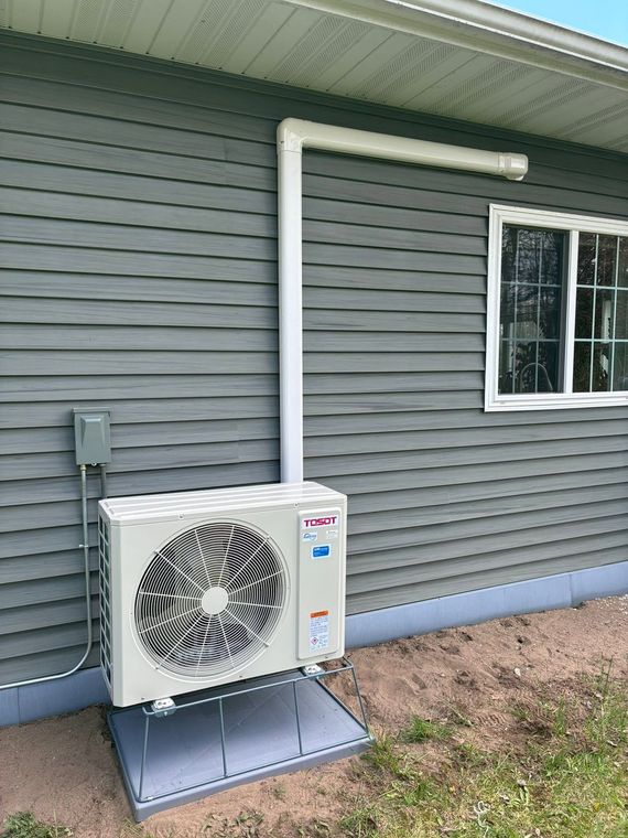 Outdoor heat pump unit mounted on a gray house wall beside a window and electrical box