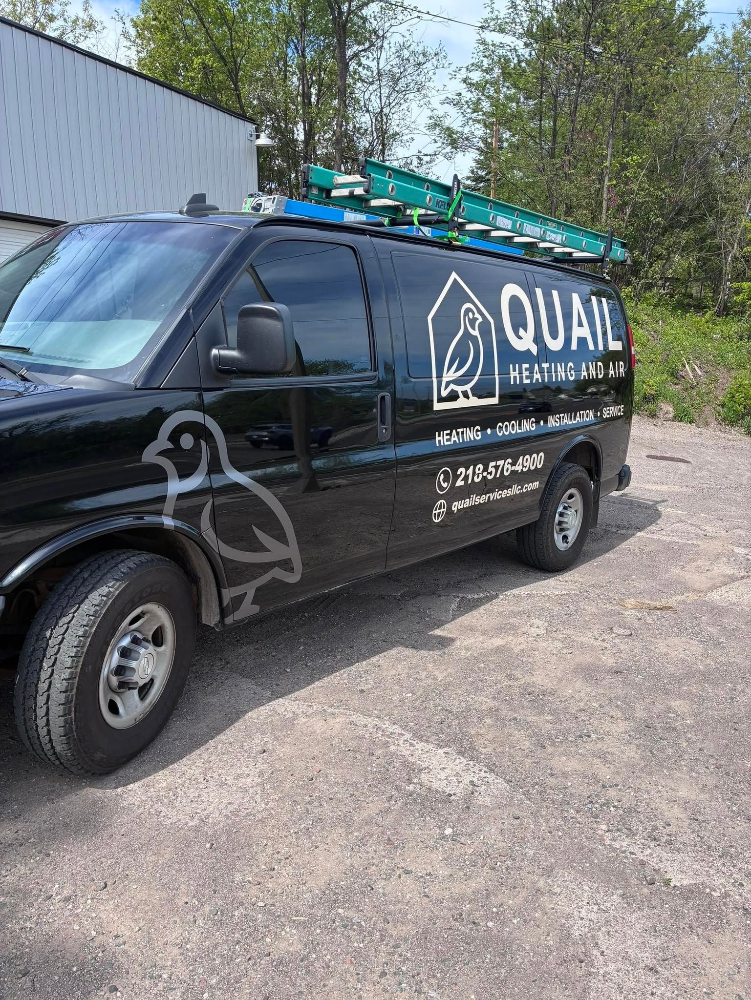 Black utility van with ladders on top parked on a gravel lot beside a building.