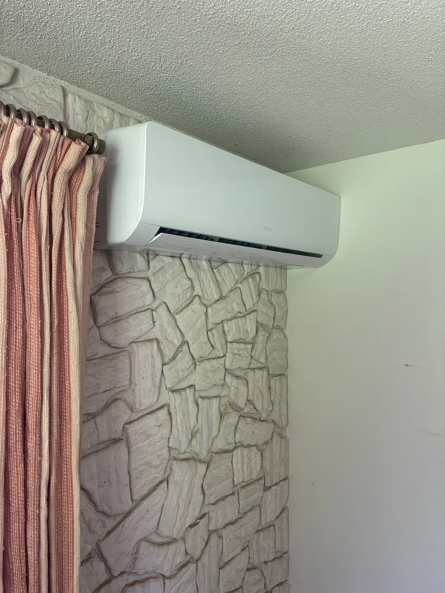 Wall-mounted white air conditioner above a stone-patterned wall beside a pink curtain