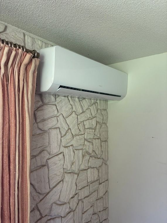 Wall-mounted air conditioner above a stone-patterned wall beside a curtain and white wall.