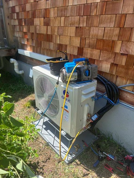 Outdoor HVAC unit by a wooden shingle wall, with tools and hoses on top in a grassy yard.