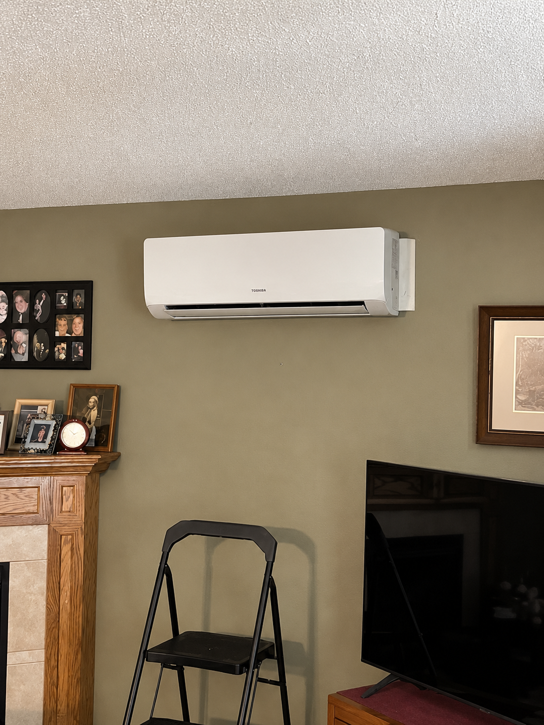 Wall-mounted white air conditioner above a TV in a living room with framed photos and a step stool