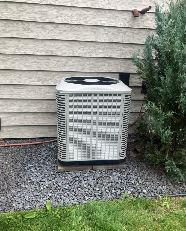 Outdoor central air conditioner beside beige house siding, near gravel and grass, partly hidden by a shrub