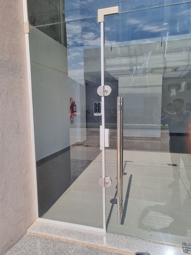 Puerta de escaparate de cristal con manilla vertical y botón pulsador de acceso junto a una pared de hormigón.