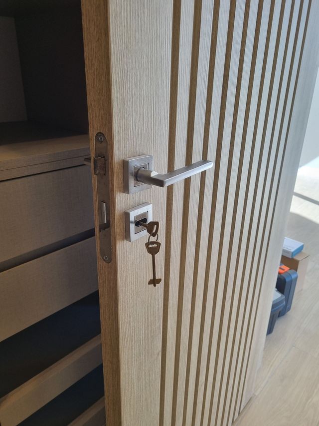 Puerta de madera abierta con manija plateada y llave insertada en la cerradura, que deja ver el interior de un armario o habitación.