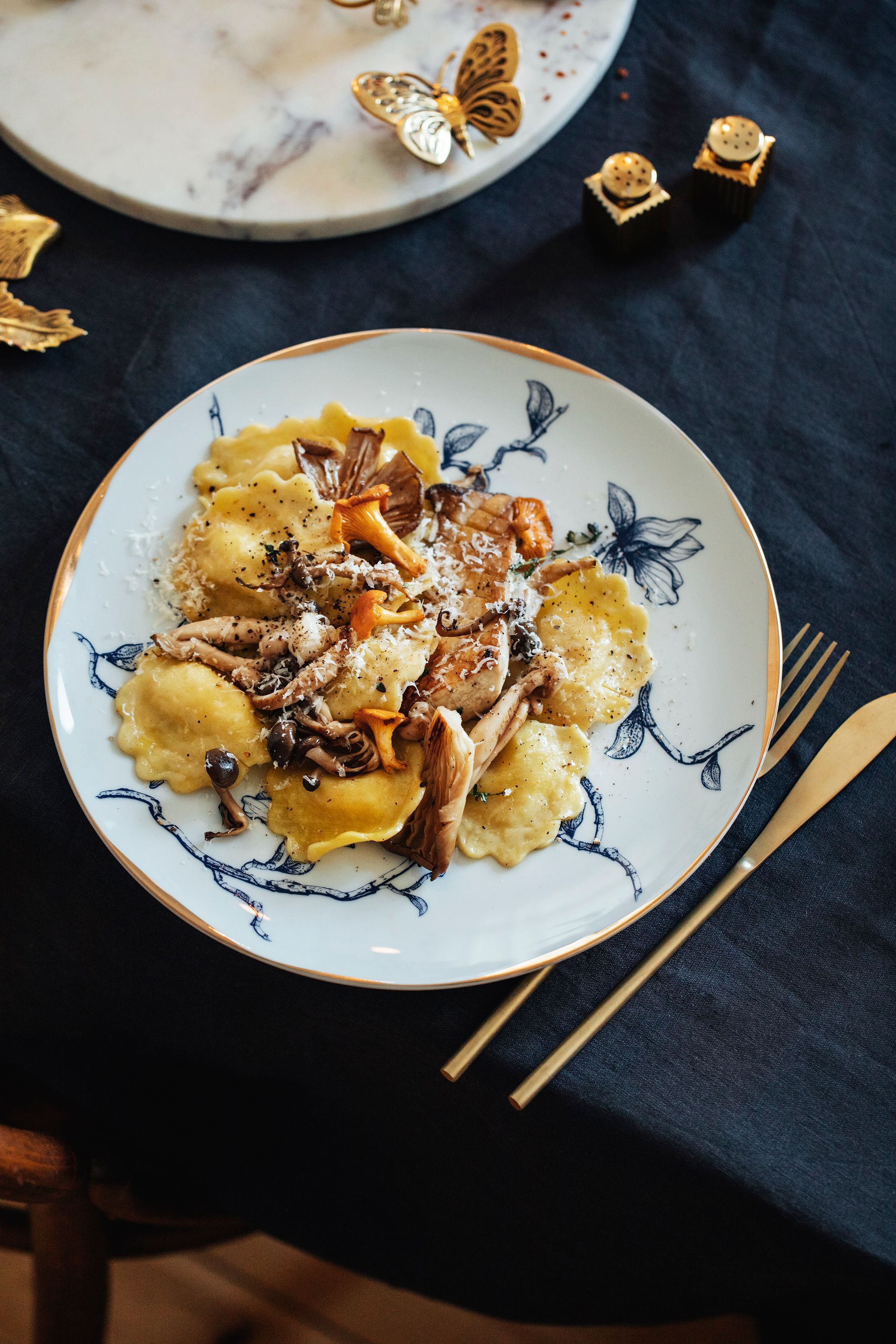 Plate of ravioli with mushrooms, garnished, on a patterned plate with gold cutlery. Food Stylist Nadine Page.