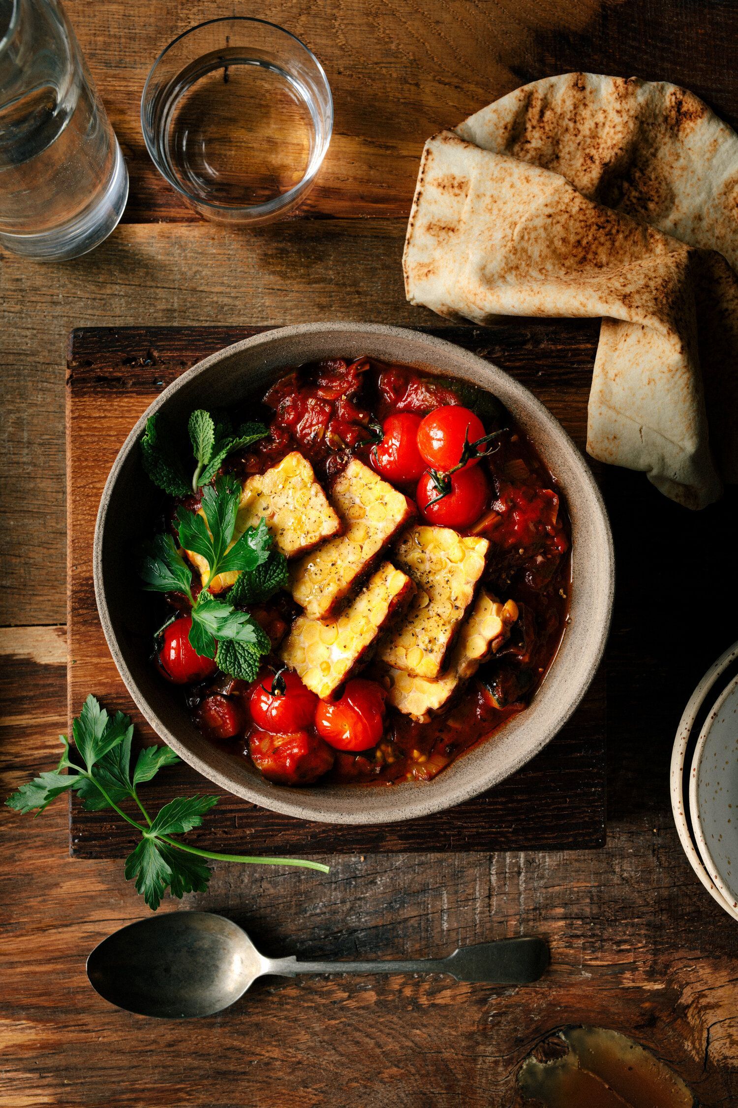 Bowl of stew with tomatoes and tempeh, garnished with parsley, with pita bread and a spoon. Food Stylist Nadine Page.