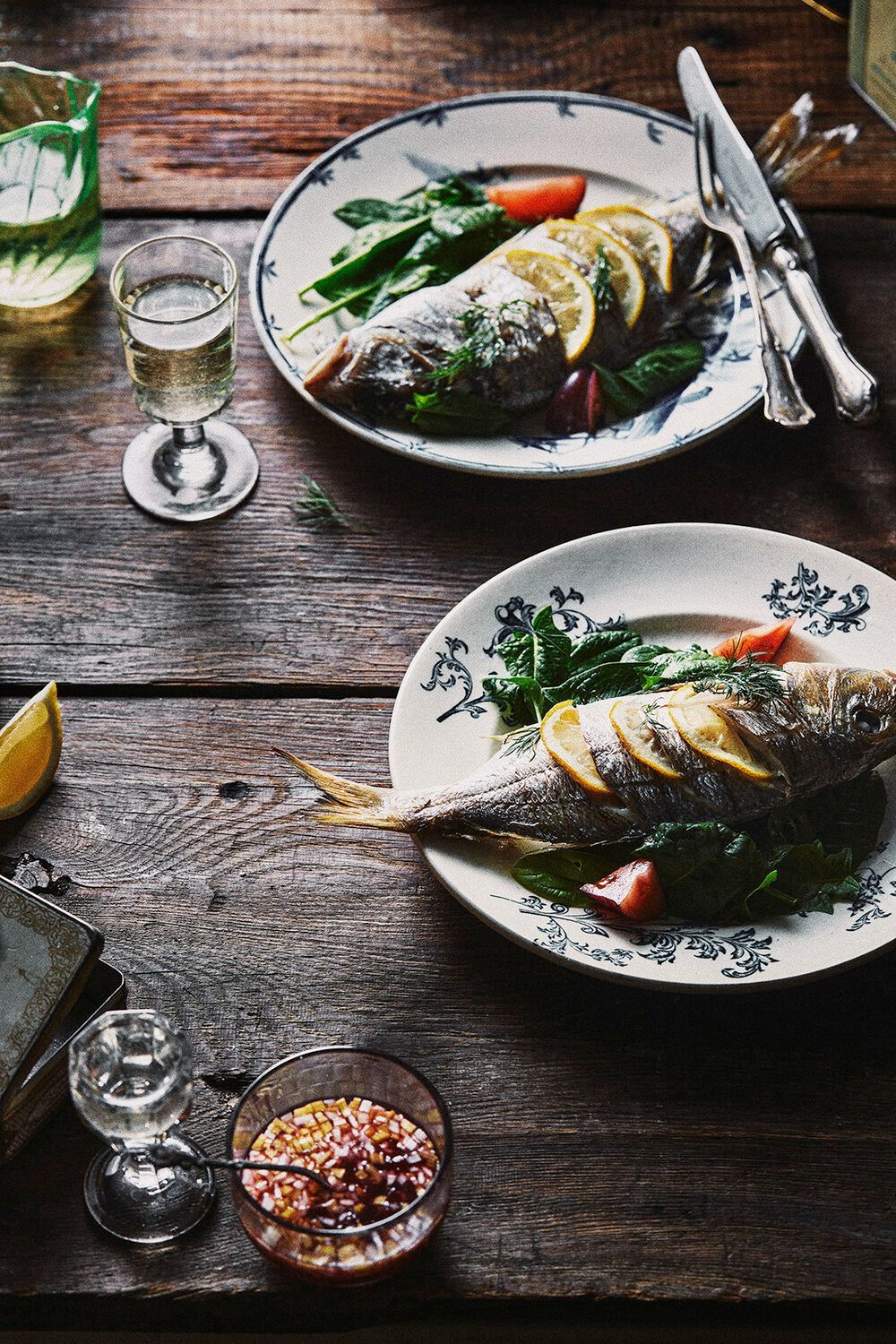 Two plates of cooked fish with lemon slices, spinach, and tomatoes on a wooden table, with drinks and silverware. Food Stylist Nadine Page.