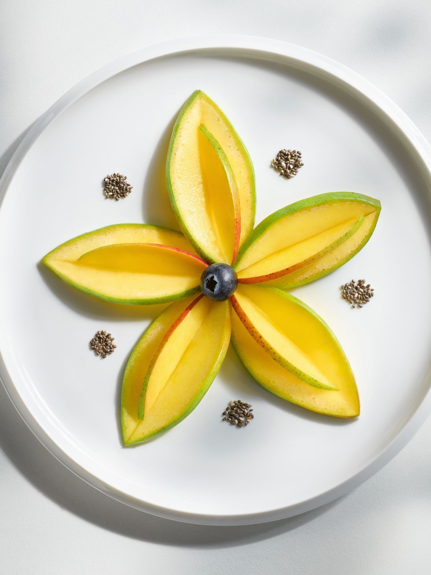Mango slices arranged in a flower shape with a blueberry center on a white plate, with seeds. Food Stylist Nadine Page.