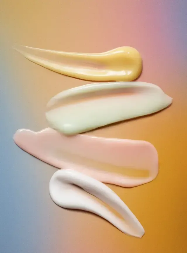 Four pastel cosmetic cream swatches in gold, white, pink, and white on a warm gradient background