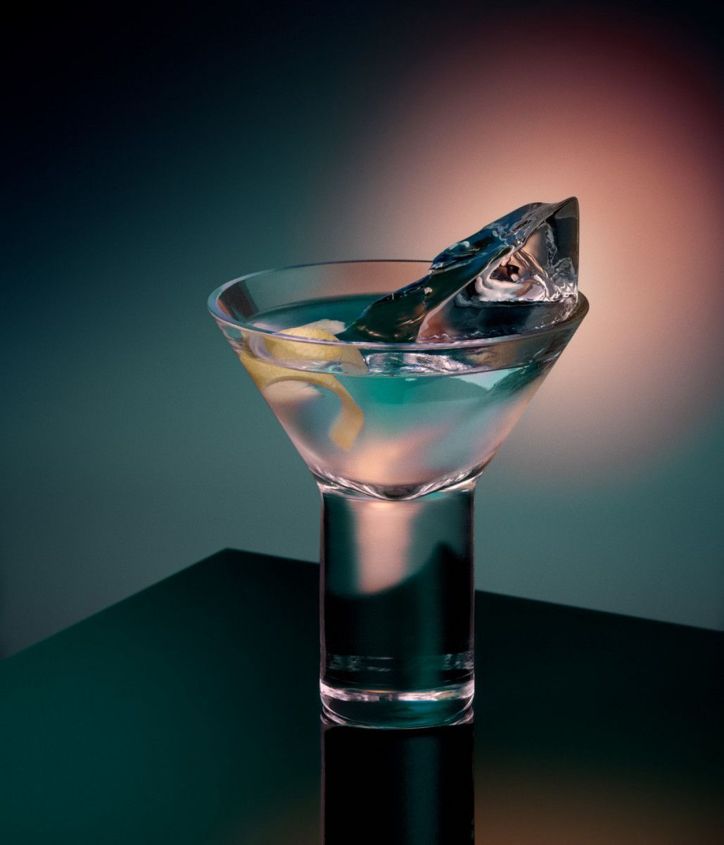 Martini glass with clear cocktail, ice, and lemon twist on a dark tabletop against a blurred background