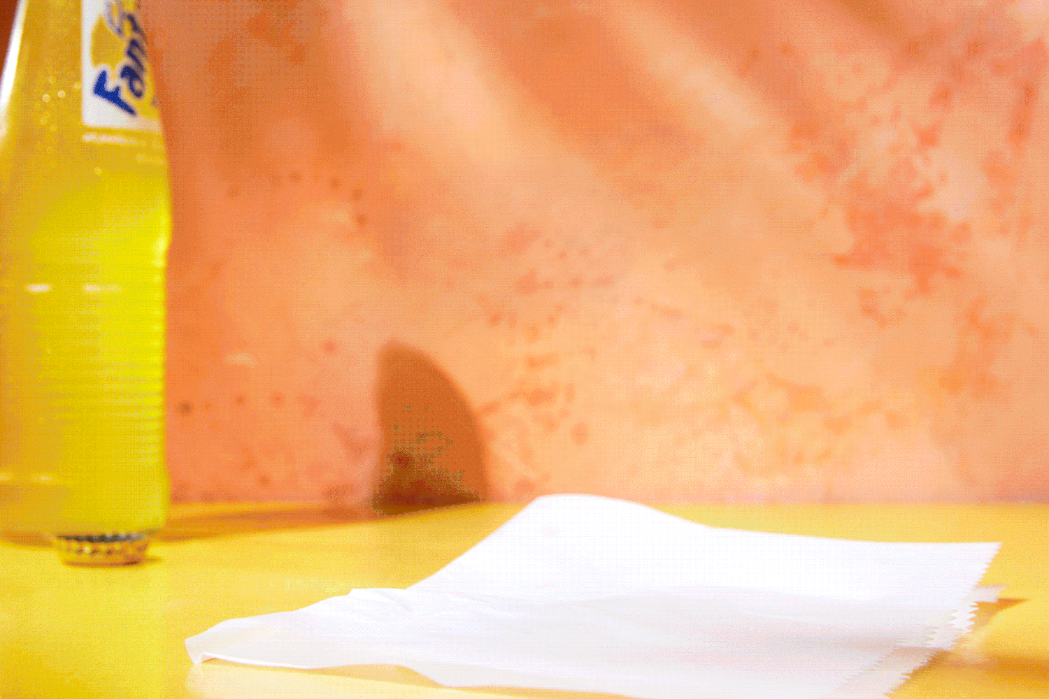 Yellow Fanta bottle on a yellow surface with a white napkin. Orange wall in the background. Food Stylist Nadine Page.