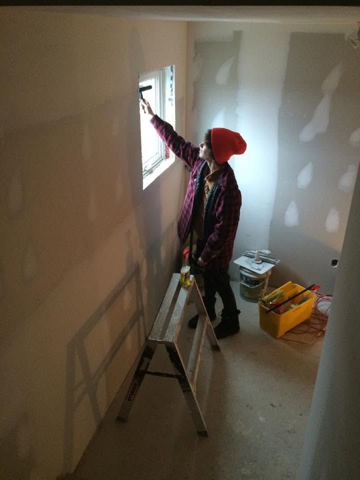 Person on ladder painting a wall near a window in an unfinished room with tools on the floor