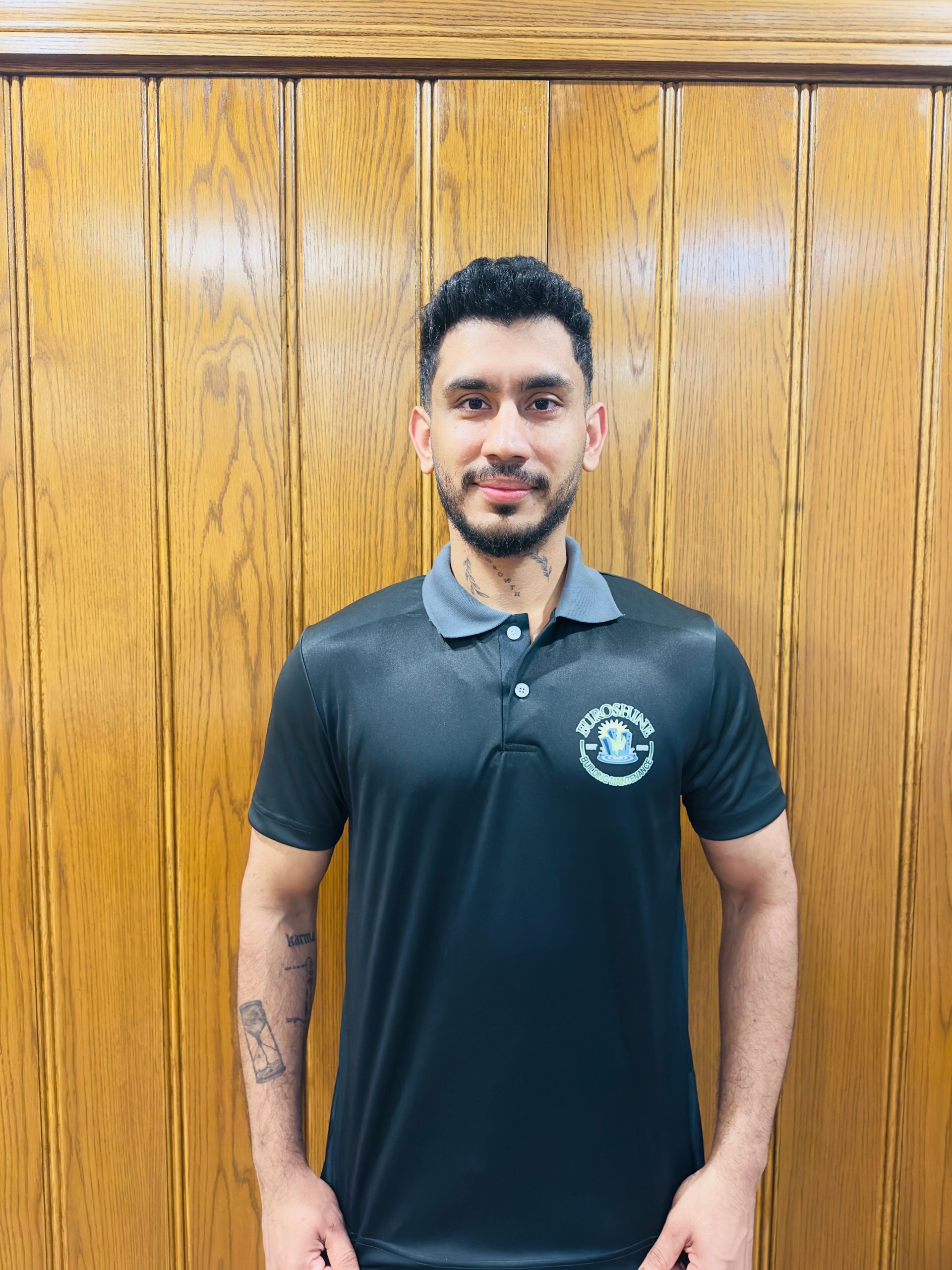 Man standing against a wooden wall, wearing a dark polo shirt with a logo, hands at his sides.
