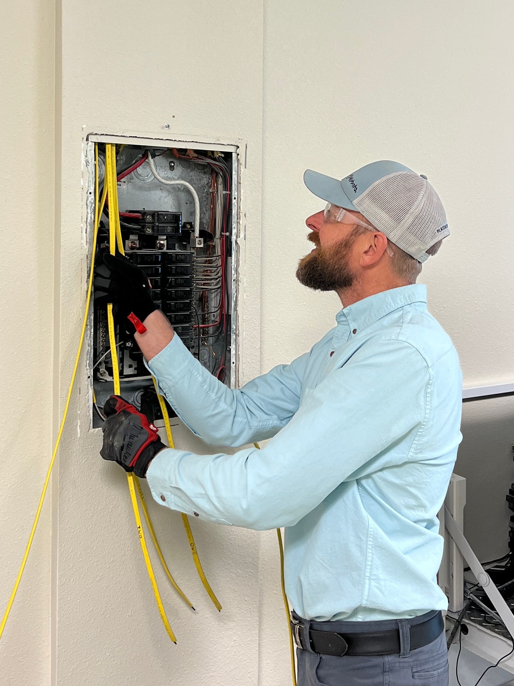 Electrician wiring an open wall panel, wearing a blue shirt, cap, and gloves.