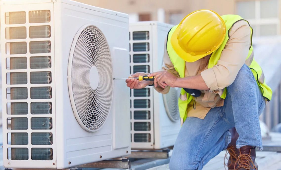 Technician in yellow hard hat repairs outdoor air conditioning unit with a screwdriver