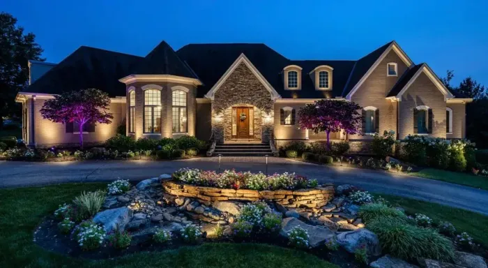 Elegant house at night, lit with exterior lights. Landscaping with illuminated water feature in front.