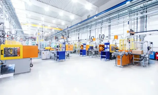 Pliers with yellow handles on top of blueprints, next to a blue pen. Factory interior with industrial machinery, orange and blue machines, white floor, bright overhead lighting.