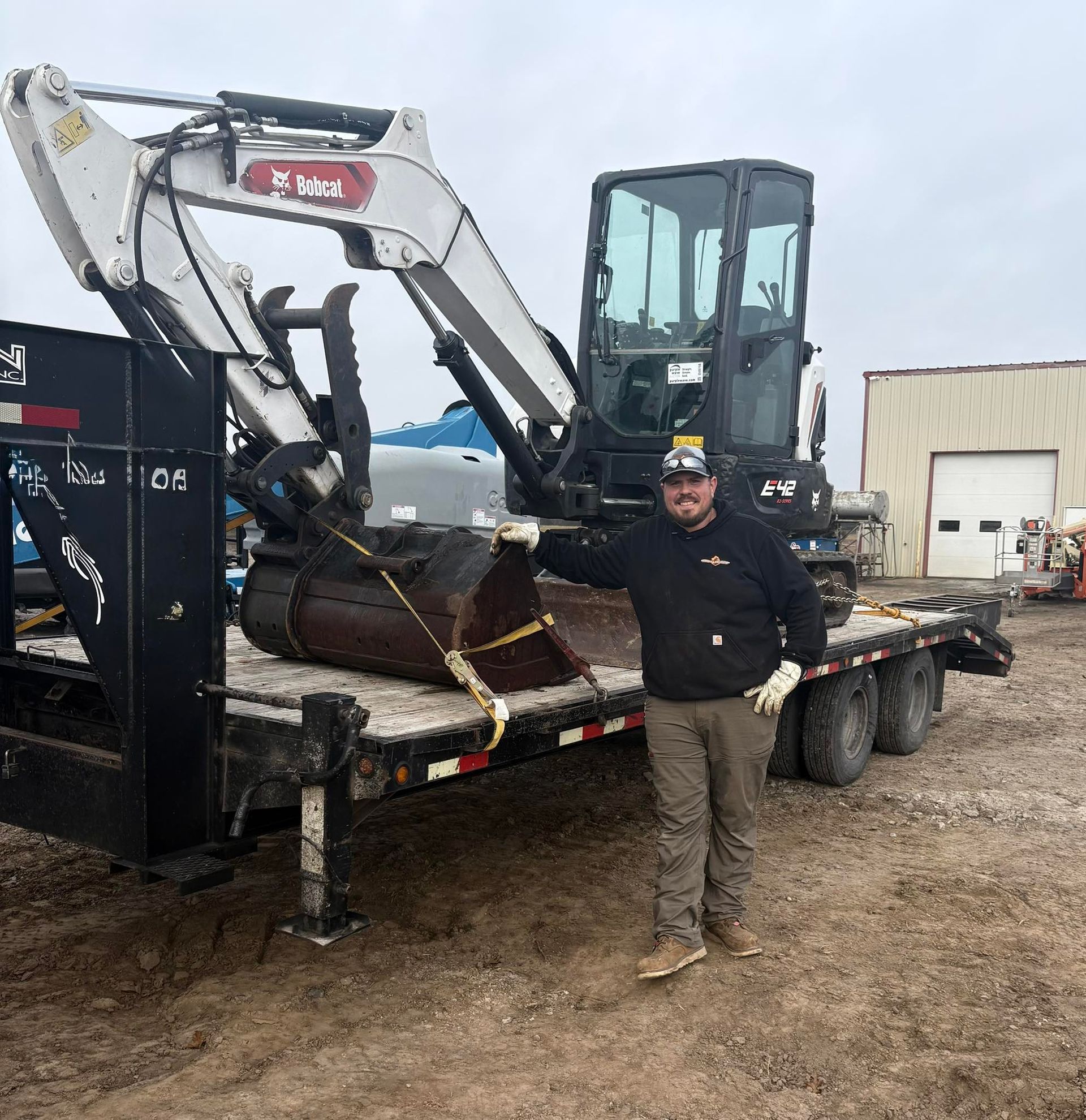 Worker standing beside a mini excavator loaded on a flatbed trailer in a muddy yard