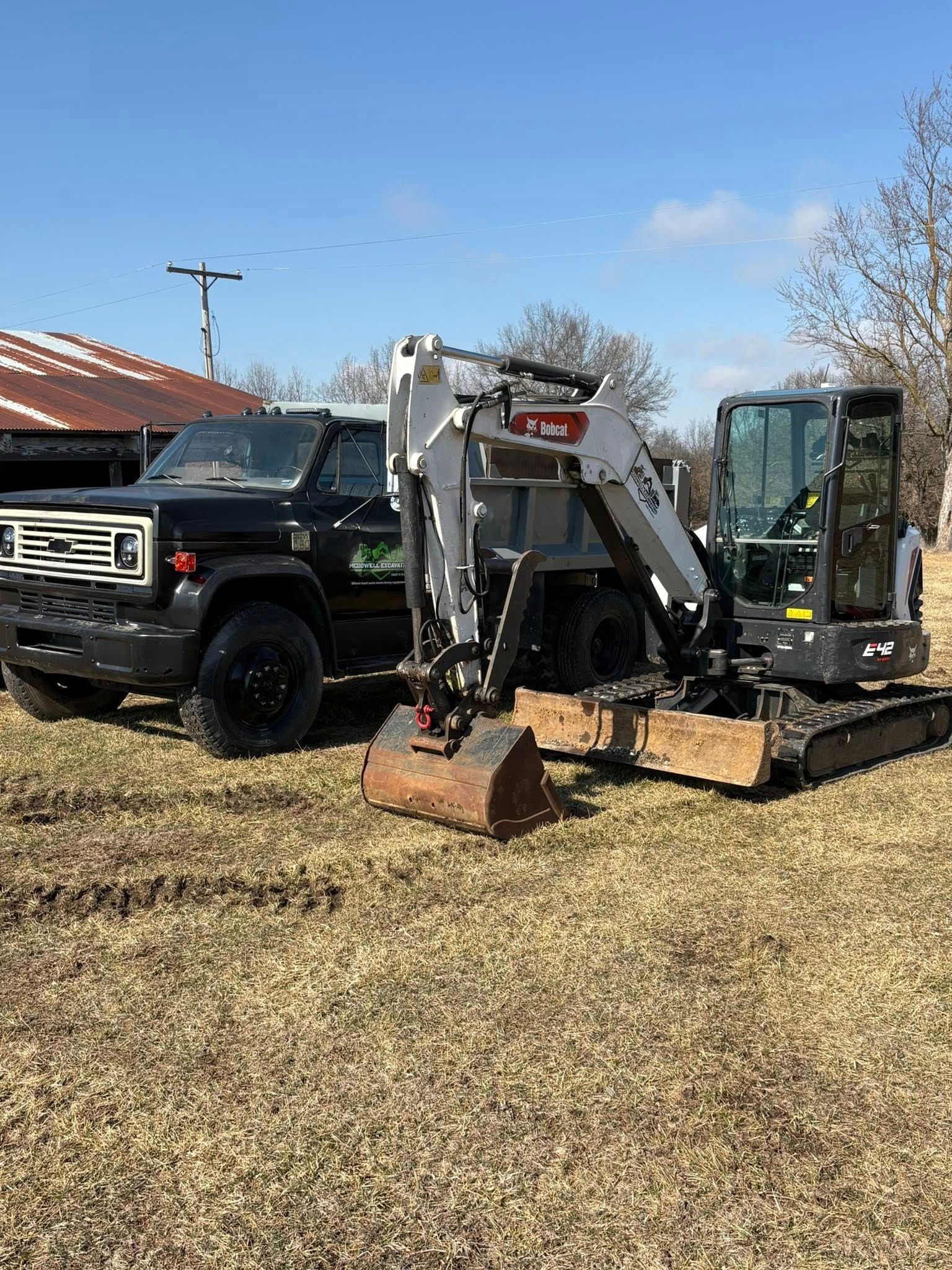 Mini excavator and black pickup truck parked on dry grass under a clear blue sky.