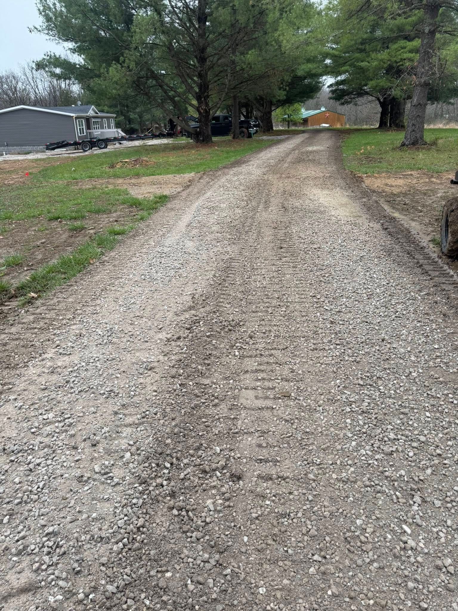 Gravel driveway leading through a wooded yard with trees and a parked RV on the left.