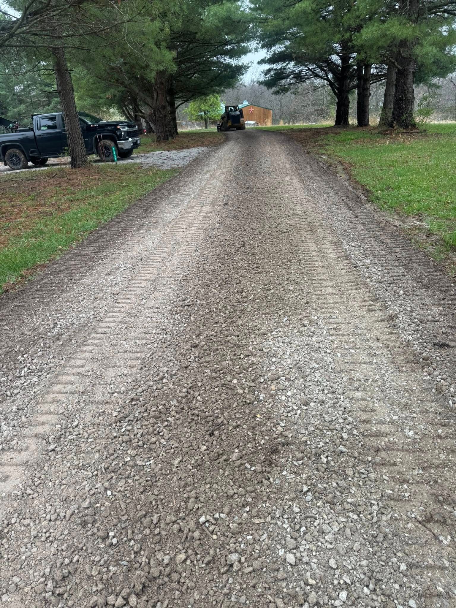 Gravel driveway lined with trees, with parked vehicles on the left and an orange truck in the distance.