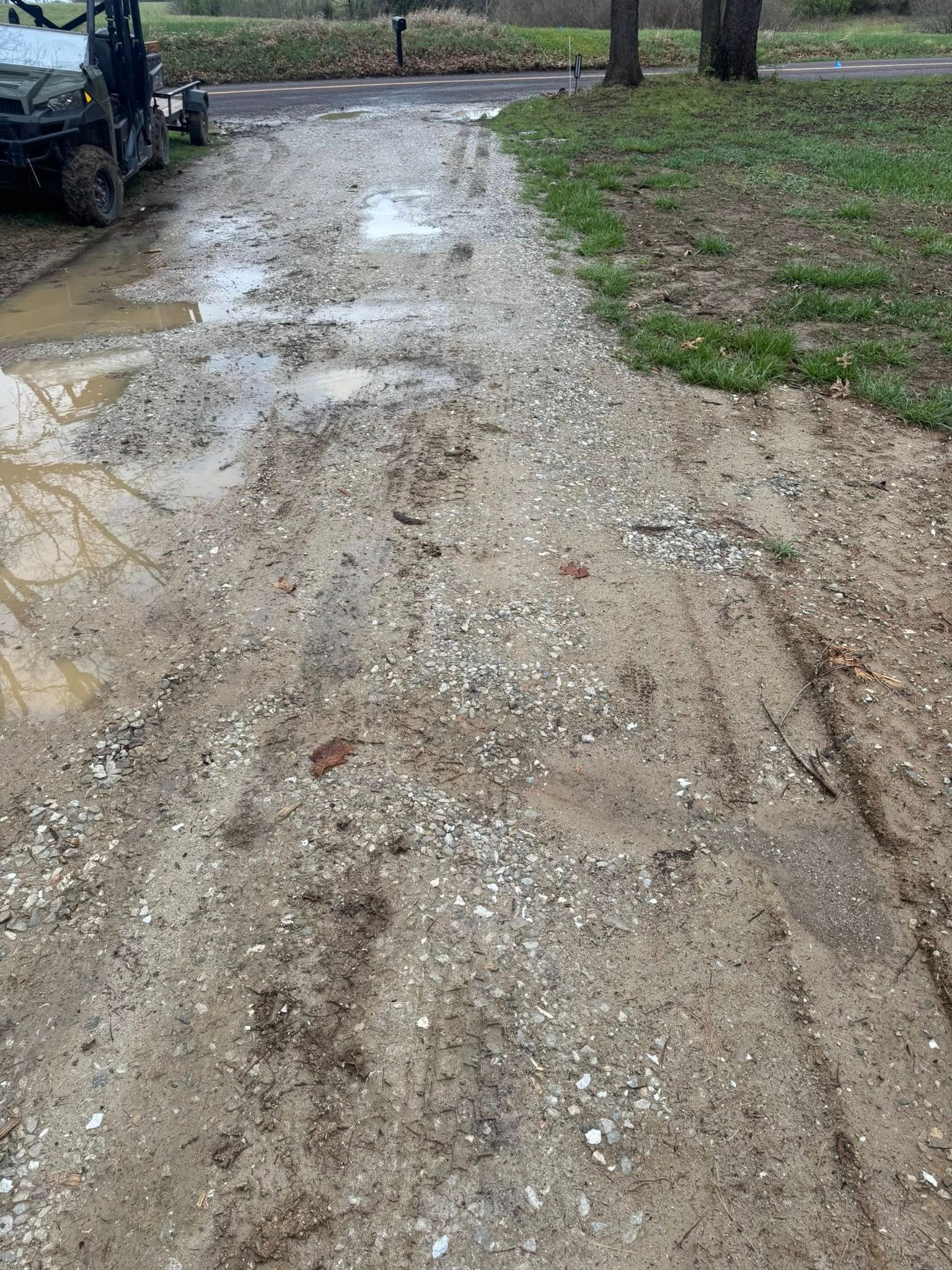 Muddy gravel driveway with puddles beside grass and a parked vehicle