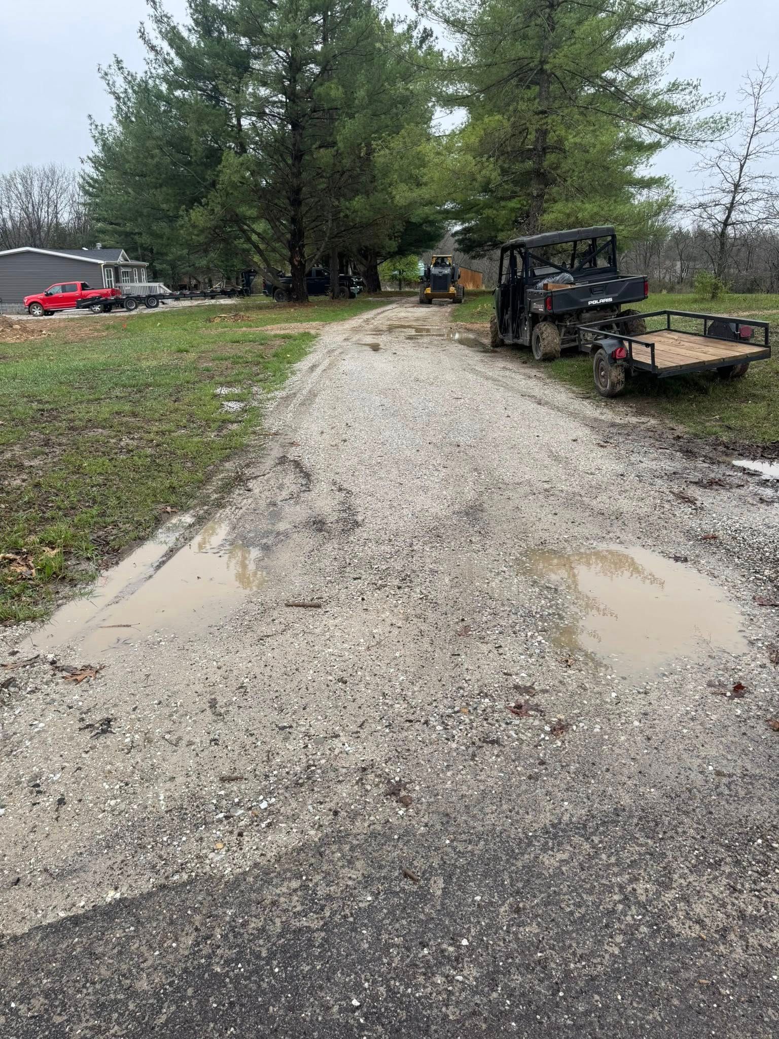 Gravel driveway with puddles beside trees, a red truck, and a utility vehicle near a house in overcast weather