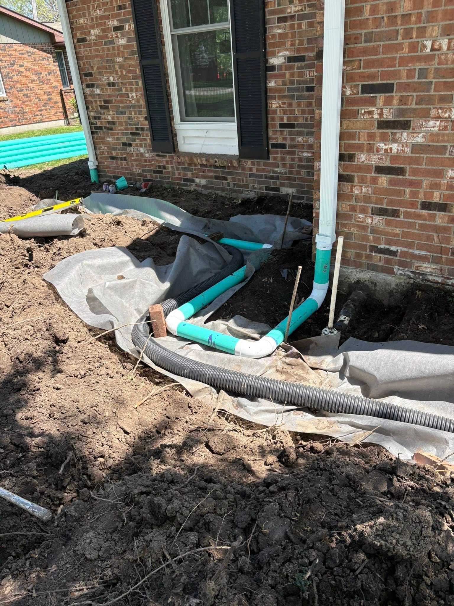 Construction trench beside brick house with exposed blue pipes and black drainage tubing over muddy soil
