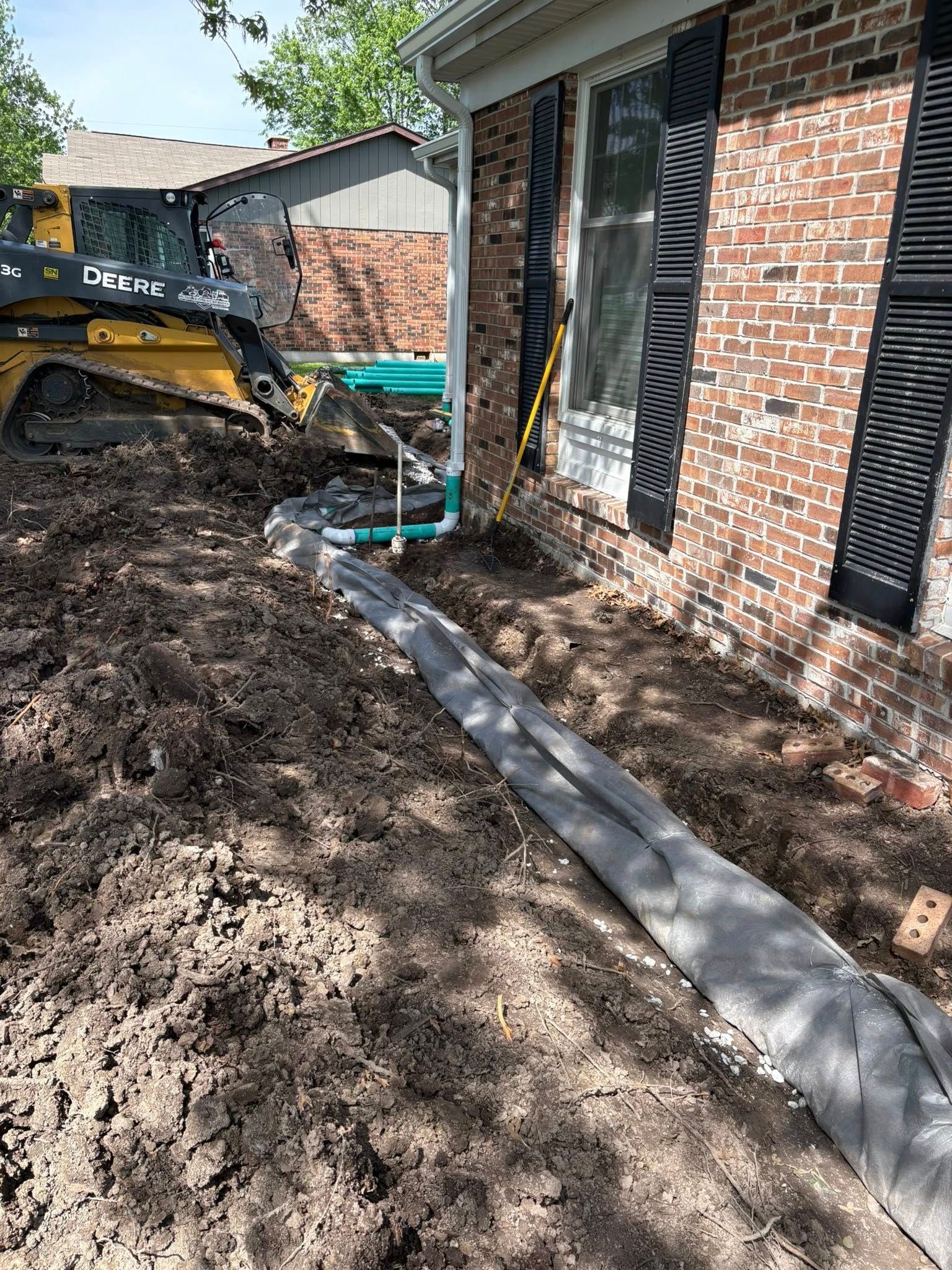 Excavator beside brick house during yard work, with dirt and a newly laid concrete border along the foundation.