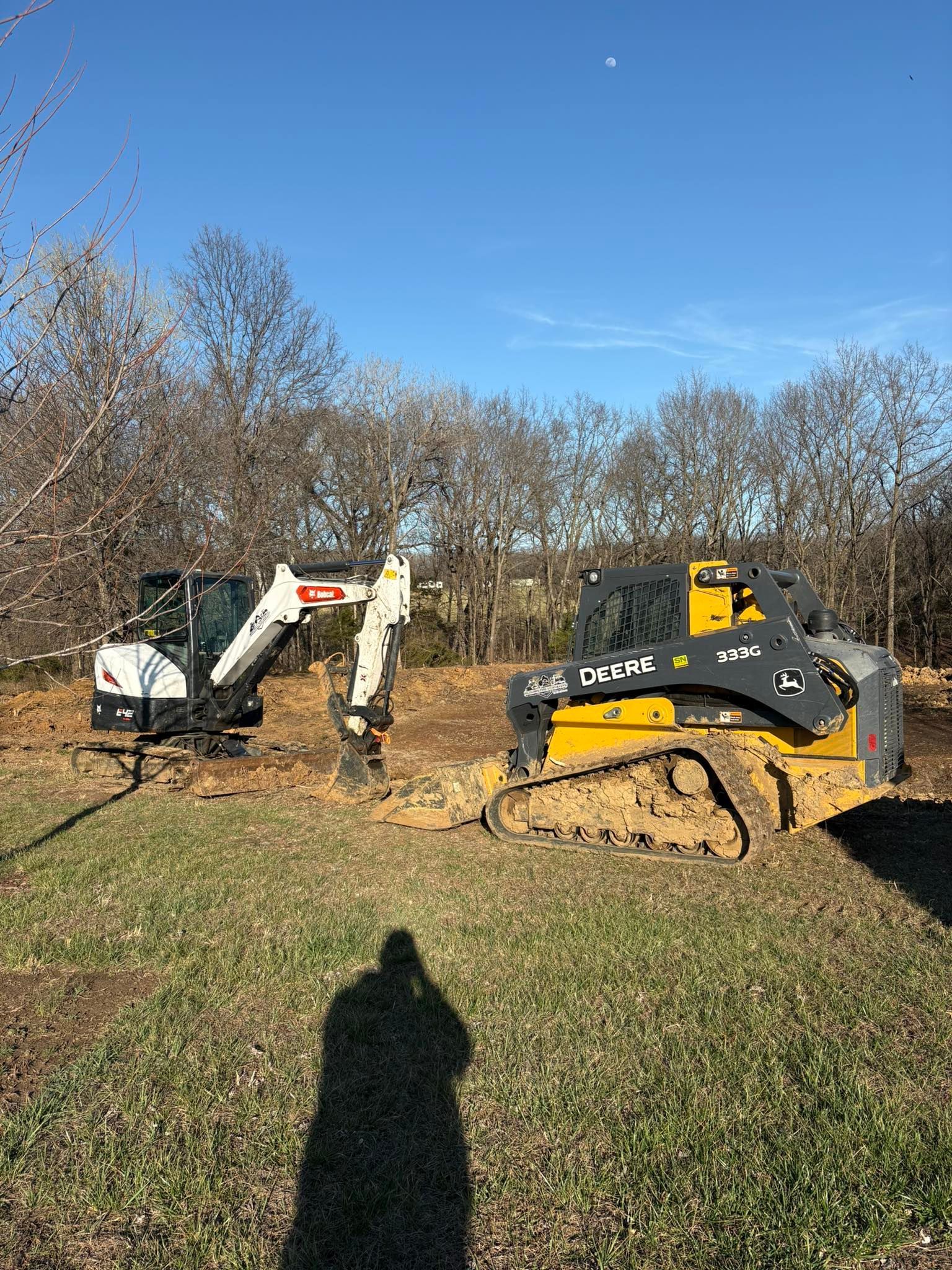 Excavator and skid steer in a grassy field with leafless trees under a clear blue sky.