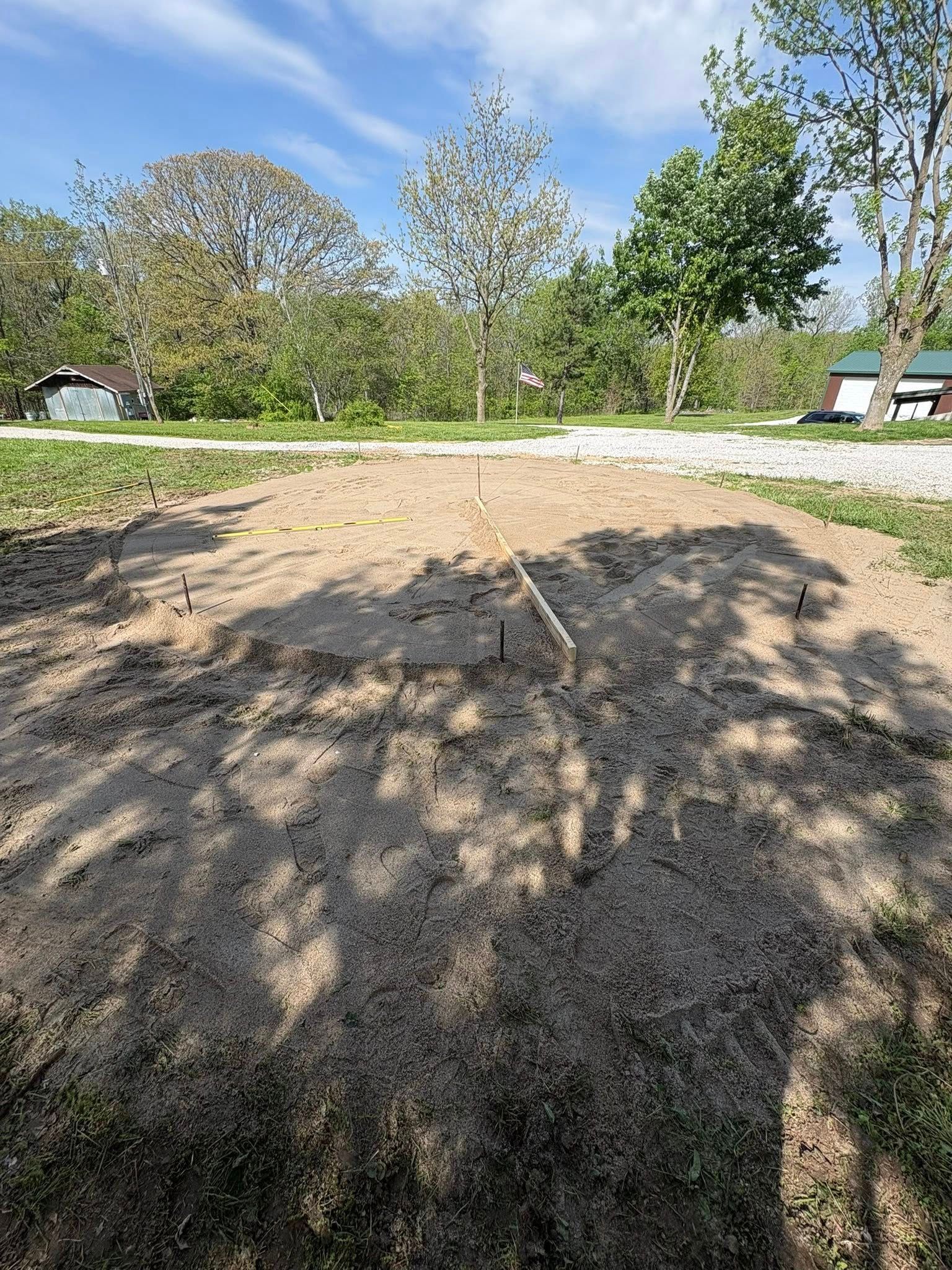 Freshly cleared dirt lot with tree shadows, grass borders, and a road with houses in the background.