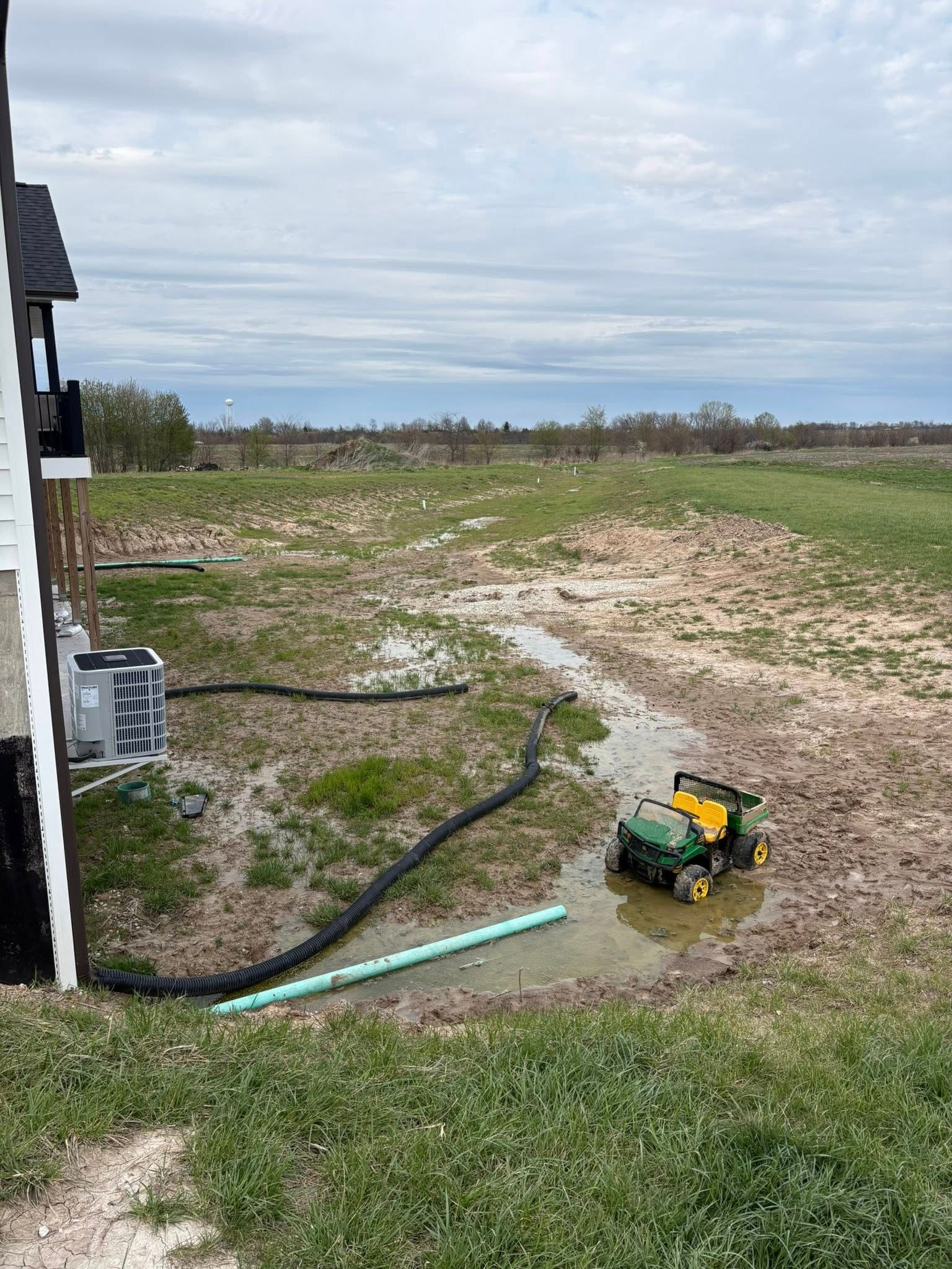 Rural yard with muddy ground, a green toy tractor, black hose, and a house edge under cloudy sky