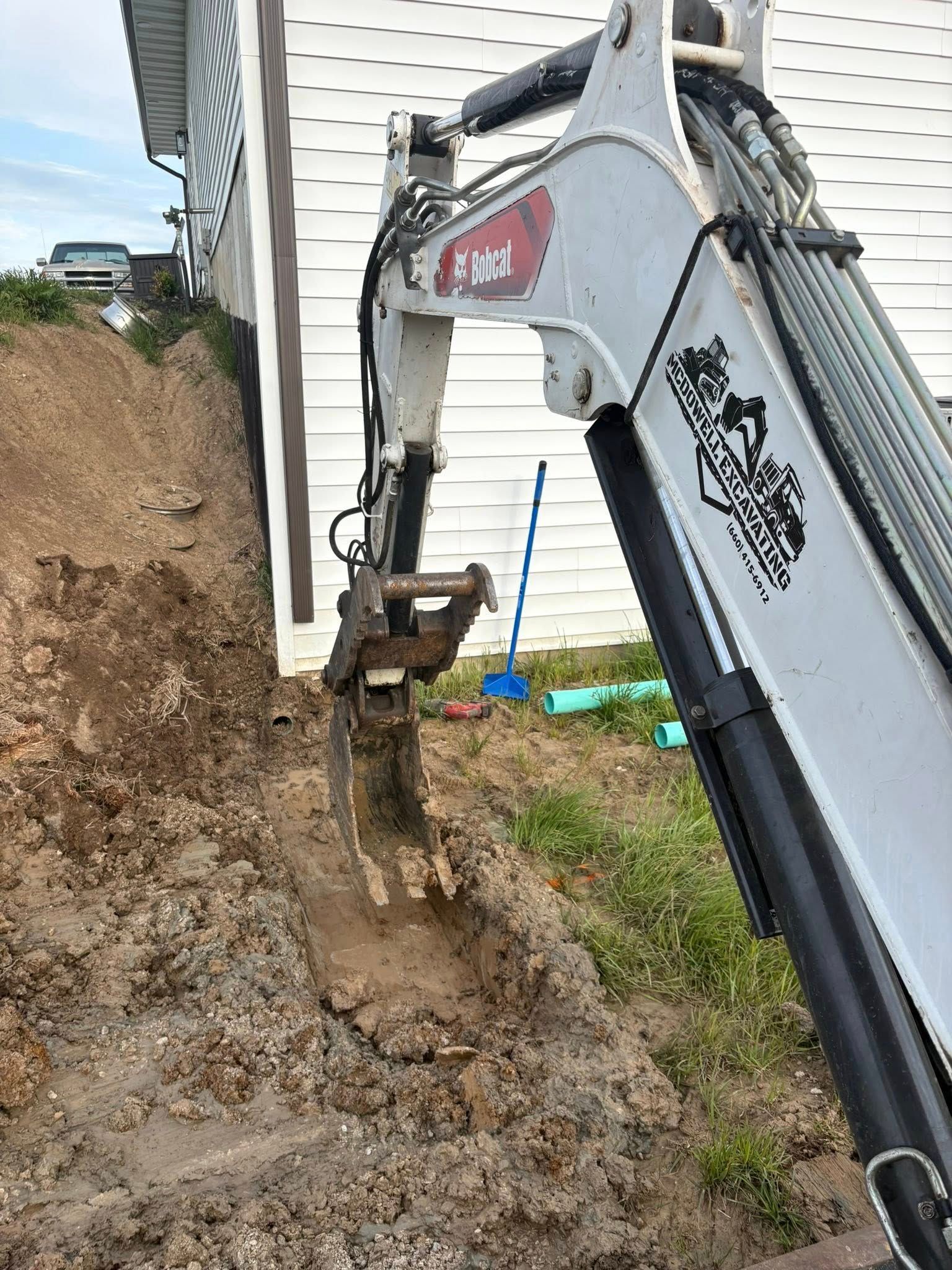 Excavator digging a muddy trench beside a white house on a steep dirt slope