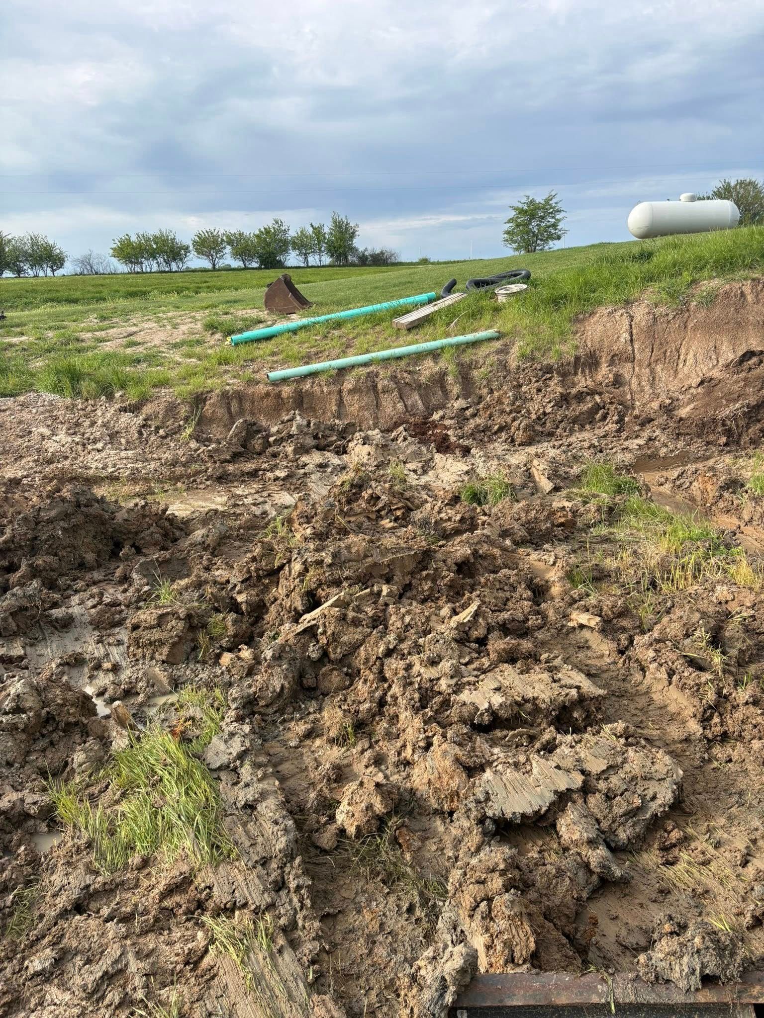 Mudslide with collapsed soil over a grassy field and a pipe in the background.