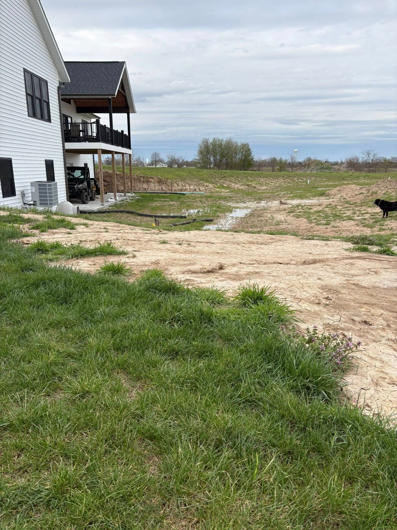 Grassy yard beside a white house with a porch, opening to a dirt path and open field under cloudy skies