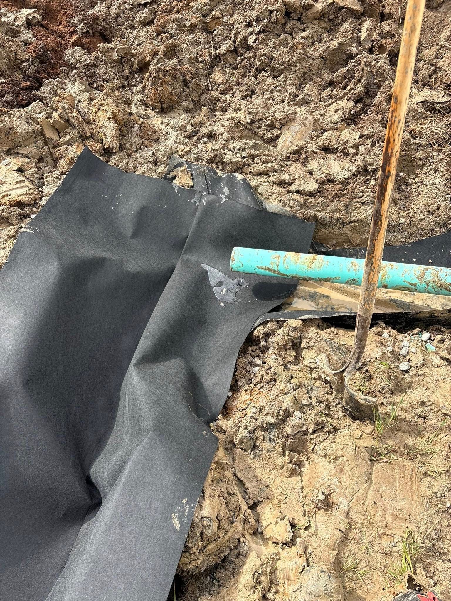 Black weed barrier fabric on soil beside a turquoise pipe and a thin wooden stake.