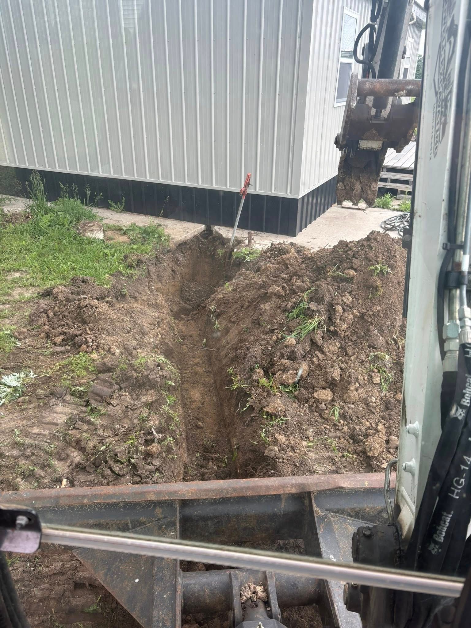 Backhoe digging a trench beside a corrugated metal fence and dirt piles in a yard