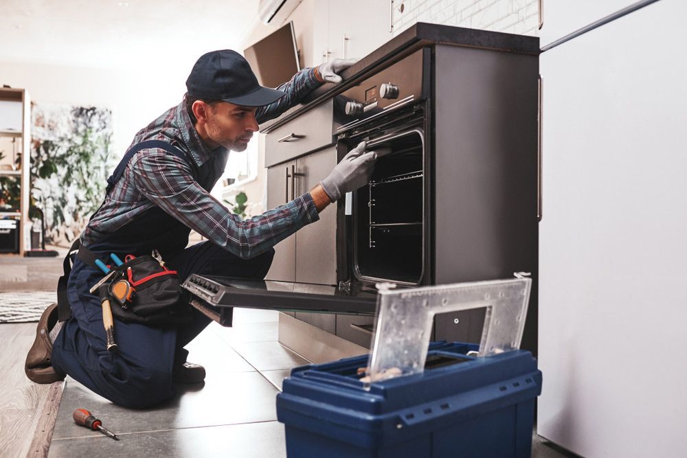 A Man is Kneeling Down in a Kitchen Fixing an Oven — Patrick Lunney Home Appliance Service & Repairs in Maudsland, QLD
