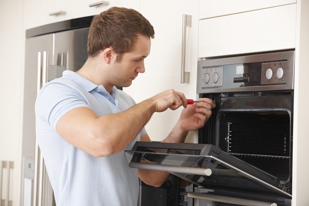 A Man is Fixing an Oven With a Screwdriver in a Kitchen — Patrick Lunney Home Appliance Service & Repairs in Maudsland, QLD