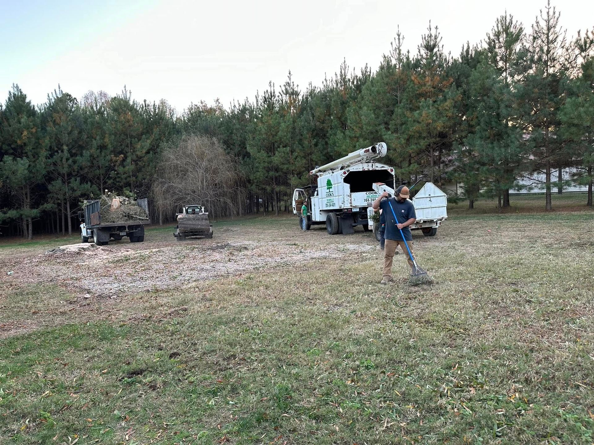 Man using a metal detector in a grassy area with tree service trucks and a treeline in the background.