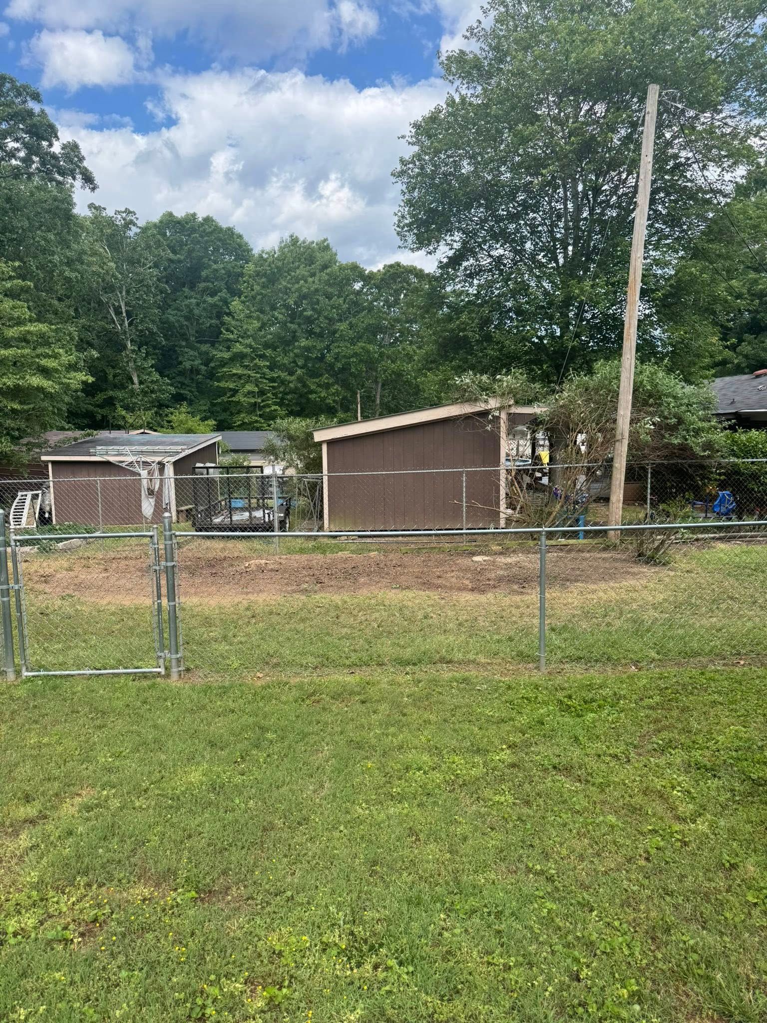 Brown sheds behind a chain link fence in a grassy yard, with a utility pole and trees in the background under a cloudy sky.