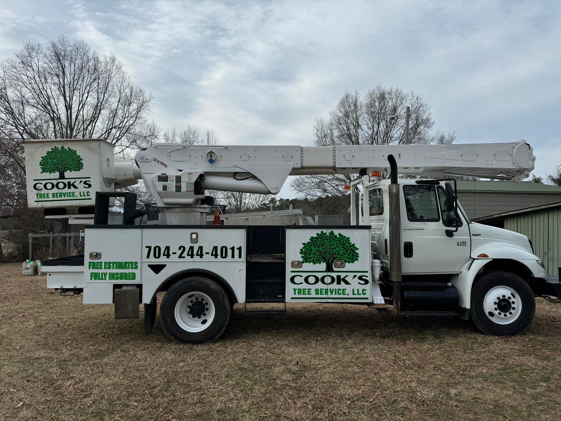 White Cook's Tree Service truck with boom extended in a yard with bare trees and a cloudy sky.