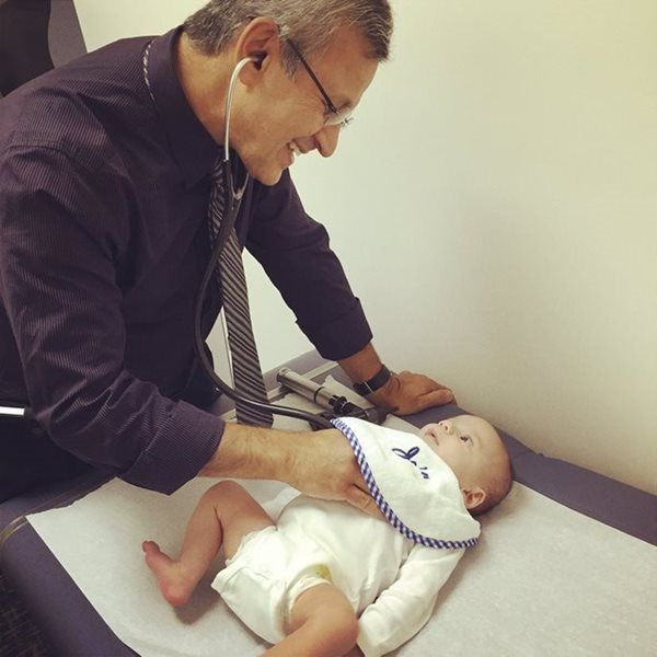 A doctor in a dress shirt and tie uses a stethoscope to examine a baby lying on an exam table.